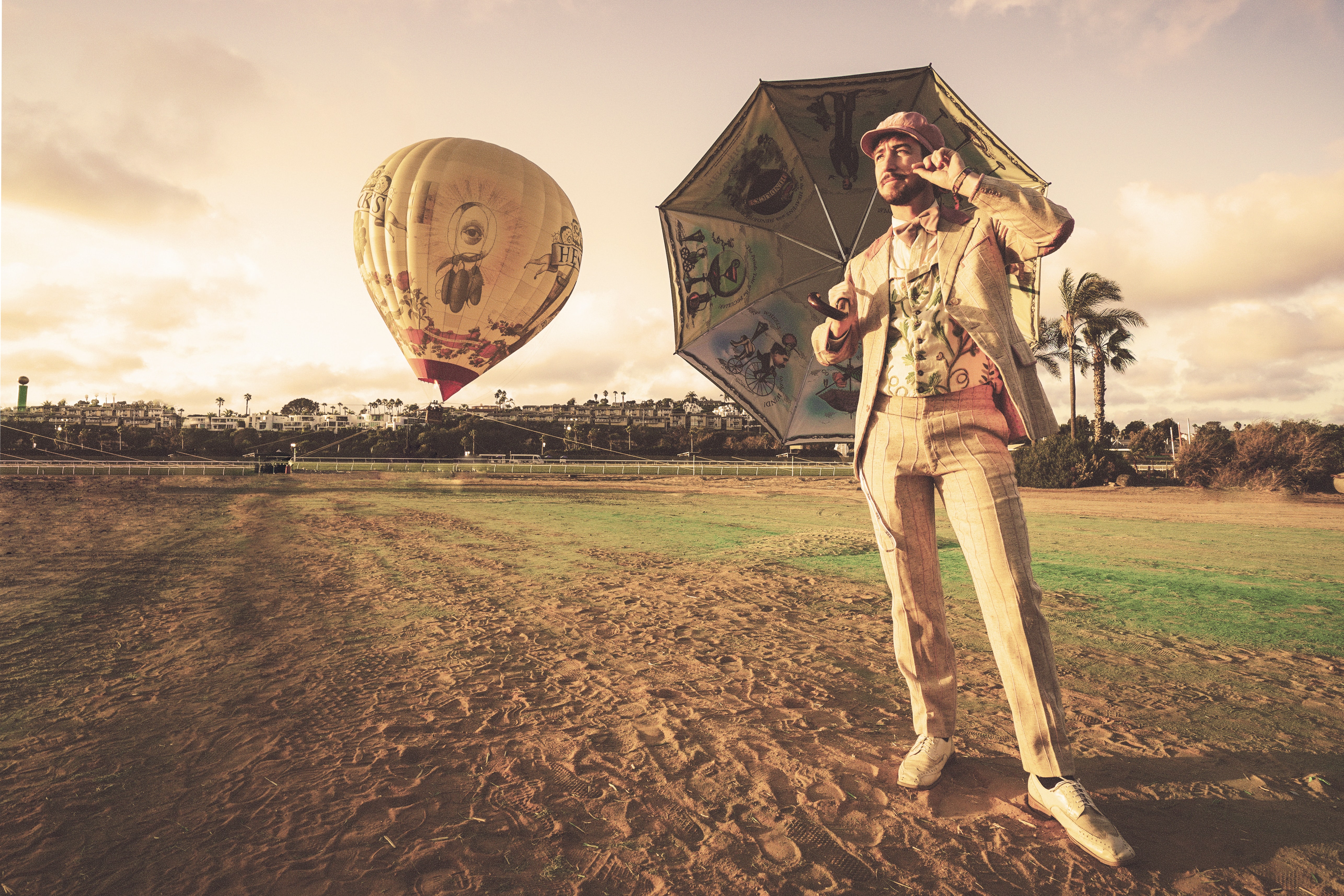 A man in a white suit stands underneath an ornate white umbrella while standing in a dirt field. A white hot air balloon hovers near the ground behind him.