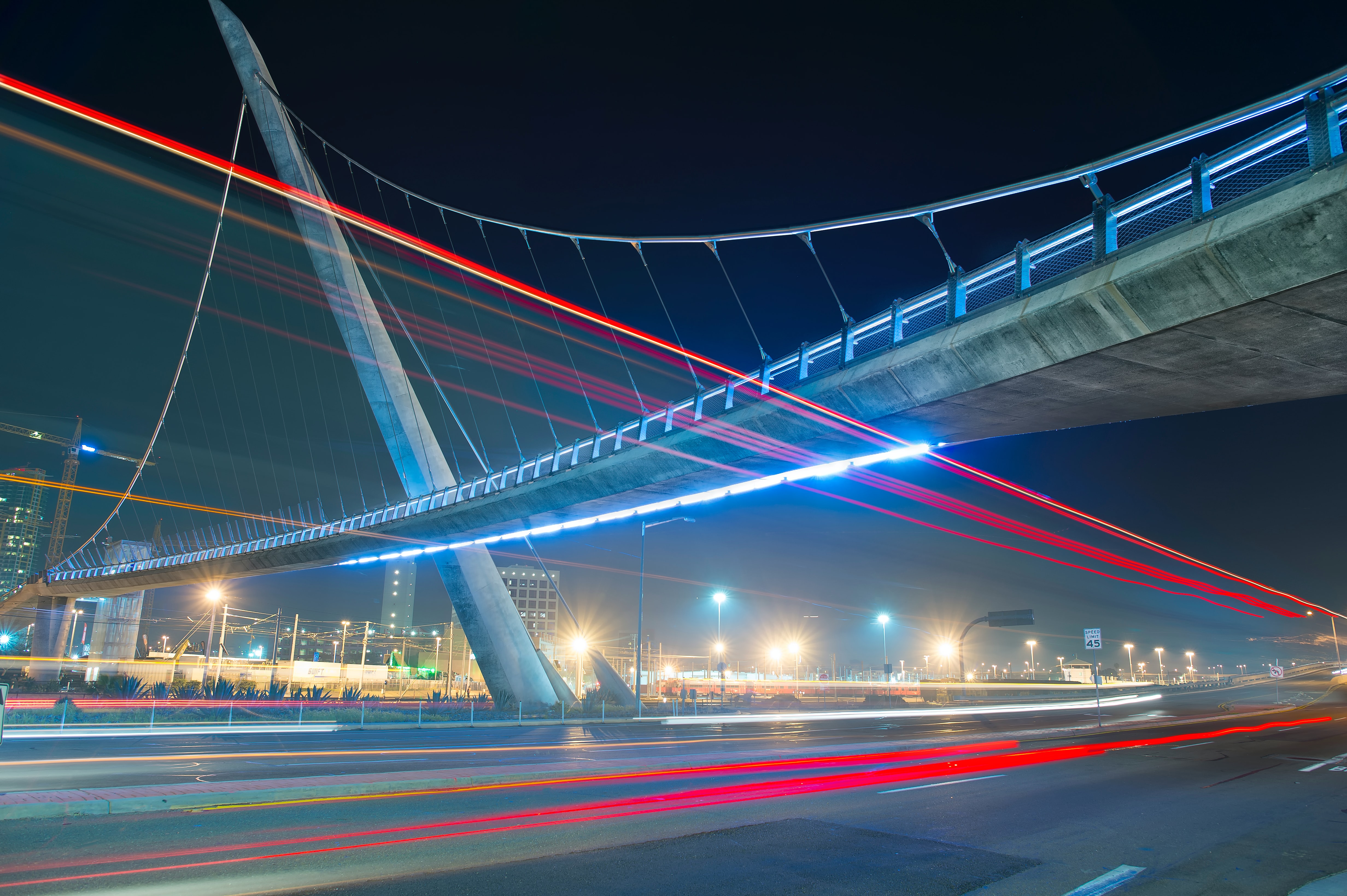 A lit pedestrian bridge in San Diego is pictured at night time. A long exposure leaves tail lights from cars streaking across the photograph's canvas.