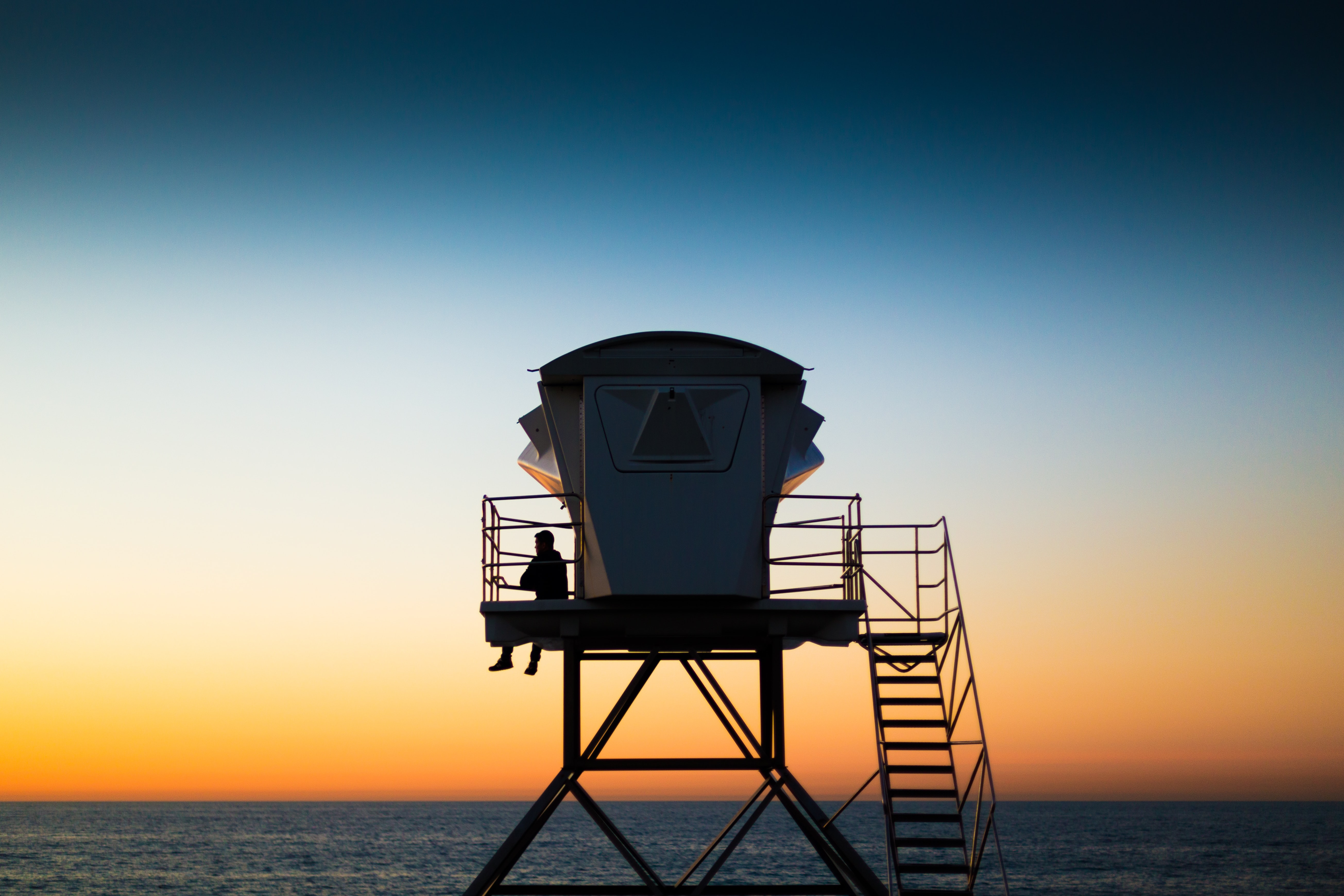 A silhouette sits atop a lifeguard tower with an orange, white and blue sunset over the ocean in the background 